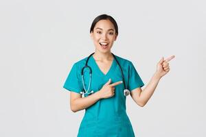 Covid-19, healthcare workers, pandemic concept. Glad smiling, beautiful asian doctor, female therapist in scrubs or nurse pointing fingers upper right corner, showing way to patient, white background photo