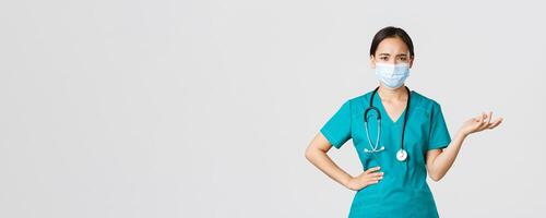 Covid-19, coronavirus disease, healthcare workers concept. Confused and upset asian female doctor in medical mask and scrubs, raise hand up puzzled, cant understand what problem, white background photo