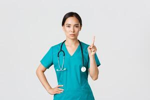 Covid-19, healthcare workers, pandemic concept. Serious-looking professional female asian doctor in scrubs, physician shaking finger in prohibition, warning gesture, forbid something photo