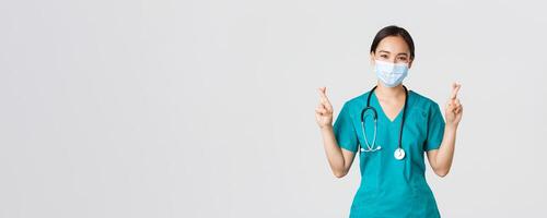 Covid-19, coronavirus disease, healthcare workers concept. Smiling optimistic asian female doctor, physician in medical mask and scrubs praying, having faith, cross fingers and make wish photo