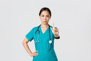 Covid-19, healthcare workers, pandemic concept. Serious-looking professional female asian doctor in scrubs, physician shaking finger in prohibition, warning gesture, forbid something photo