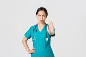 Covid-19, healthcare workers, pandemic concept. Angry serious-looking asian doctor, female physician or nurse in scrubs frowning displeased, extend hand to show stop, disagree, prohibit or forbid photo