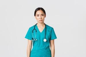 COVID-19, cuidado de la salud trabajadores, pandemia concepto. agotado joven asiático hembra enfermero, médico mirando cansado después cambio en hospital, mirando triste con fatiga, en pie blanco antecedentes en matorrales foto