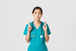 Covid-19, healthcare workers, pandemic concept. Hopeful nervous asian female intern in scrubs waiting for news, doctor cross fingers and biting lip as looking upper left corner, praying photo