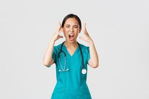 Covid-19, healthcare workers and preventing virus concept. Annoyed and angry, pissed-off asian doctor, female nurse in scrubs going mad, screaming in rage, shaking hands outraged, white background photo