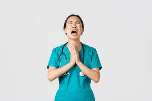 Covid-19, healthcare workers, pandemic concept. Overworked sad and tired asian female doctor, nurse begging for help, pleading god and crying from stress, standing white background photo