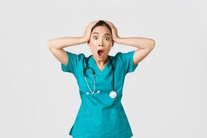 Covid-19, healthcare workers and preventing virus concept. Shocked and anxious asian female doctor in panic, grab head and screaming alarmed, have big problem, got in trouble, white background photo