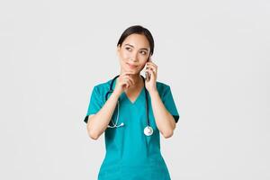 COVID-19, cuidado de la salud trabajadores y prevenir virus concepto. retrato de sonriente asiático hembra doctor, interno en matorrales hablando en teléfono y mirando considerado, pensando o haciendo elección foto