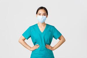 Covid-19, coronavirus disease, healthcare workers concept. Cheerful smiling asian female doctor, physician in scrubs and medical mask, looking upbeat, working with patients in hospital photo