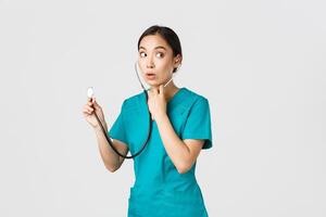 Covid-19, healthcare workers and preventing virus concept. Intrigued asian female doctor, physician in scrubs, looking away thoughtful, listening to patient lungs with stethoscope, white background photo