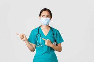 Covid-19, coronavirus disease, healthcare workers concept. Skeptical and disappointed asian female doctor, nurse in scrubs and medical mask looking displeased, cringe aversion, pointing left photo