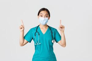 Covid-19, coronavirus disease, healthcare workers concept. Intrigued asian female doctor, physician or intern in medical mask and scrubs, looking curious, pointing fingers up, white background photo