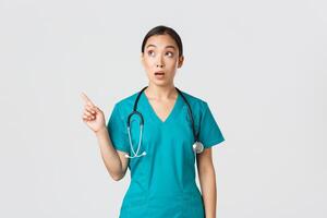 Covid-19, healthcare workers, pandemic concept. Surprised and amazed female asian physician looking and pointing upper left corner. Astonished korean doctor stare at banner with advertisement photo