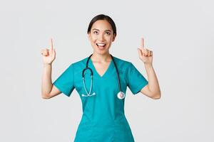 Covid-19, healthcare workers, pandemic concept. Amused cheerful asian female nurse, doctor or surgeon in scrubs pointing fingers and looking up with satisfied face, standing white background photo