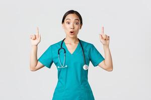 Covid-19, healthcare workers, pandemic concept. Surprised and thrilled asian female nurse, woman doctor in scrubs asking question interesting promo, pointing fingers up, showing clinic advertisement photo