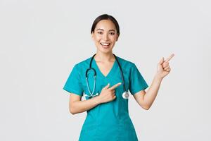 Covid-19, healthcare workers, pandemic concept. Smiling cheerful asian female doctor, therapist in scrubs pointing fingers upper right corner, inviting for checkup, recommend vaccinate photo