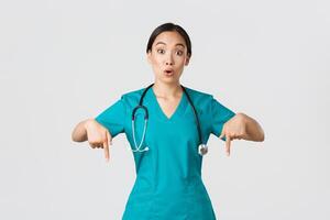 Covid-19, healthcare workers, pandemic concept. Amazed and impressed asian female nurse, doctor in scrubs pointing fingers down and looking questioned, interested in new promo, white background photo