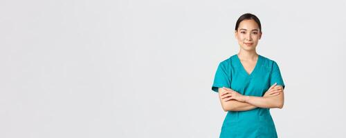 Covid-19, healthcare workers, pandemic concept. Confident smiling asian nurse in scrubs standing self-assured, cross arms chest. Professional doctor ready for surgery, standing white background photo