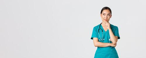 Covid-19, healthcare workers, pandemic concept. Thoughtful smart asian nurse in scrubs looking away and thinking, smiling pleased. Doctor have interesting idea, pondering over white background photo