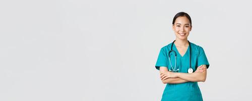 Covid-19, healthcare workers, pandemic concept. Portrait of confident smiling, attractive asian female nurse in scrubs, with stethoscope, cross arms chest and looking at camera, white background photo