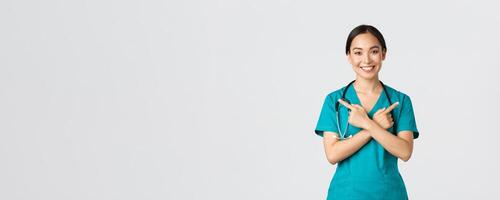 Covid-19, healthcare workers, pandemic concept. Smiling beautiful asian nurse, intern or doctor in scrubs pointing sideways and looking at camera, showing variants, two choices, white background photo