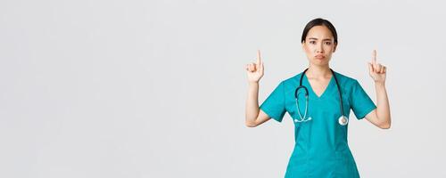 Covid-19, healthcare workers, pandemic concept. Gloomy disappointed asian female doctor, physician in scrubs pointing fingers up and grimacing displeased, standing white background photo