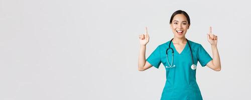 Covid-19, healthcare workers, pandemic concept. Smiling cheerful pretty nurse, female doctor or intern in scrubs pointing fingers up, showing banner, make announcement, demonstrate advertisement photo