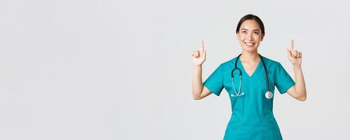 COVID-19, cuidado de la salud trabajadores, pandemia concepto. sonriente hermosa asiático hembra enfermero, médico en matorrales señalando y mirando arriba con satisfecho sonrisa, contento a espectáculo increíble promoción oferta, blanco antecedentes foto