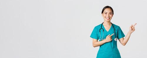 Covid-19, healthcare workers, pandemic concept. Smiling beautiful asian female nurse, intern pointing fingers upper right corner, inviting for checkup, online clinic appointment, white background photo