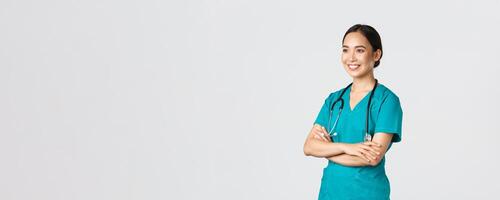 Covid-19, healthcare workers, pandemic concept. Side view of professional confident and hopeful asian female doctor, nurse looking assured away and smiling, standing in scrubs white background photo