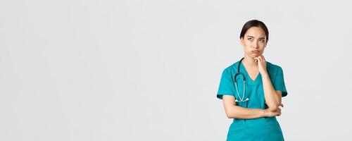 Covid-19, healthcare workers, pandemic concept. Annoyed and bothered asian female doctor, nurse on her shift looking irritated or tired, eye roll bored and exhale, standing white background in scrubs photo