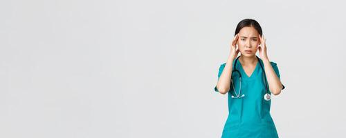 Covid-19, healthcare workers, pandemic concept. Overworked and exhausted asian female doctor, physician feeling sick, wear scrubs, touching head, complaining on headache or high fever photo
