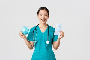 Covid-19, coronavirus disease, healthcare workers concept. Amused smiling asian female nurse, inters in scrubs showing respirator and medical mask, receive personal protective equipment photo