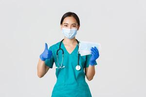 Covid-19, coronavirus disease, healthcare workers concept. Smiling asian doctor, nurse in scrubs and rubber gloves, showing thumbs-up and medical masks, white background photo