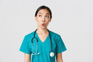 Healthcare workers, preventing virus, quarantine campaign concept. Impressed and amazed asian female doctor, nurse in medical scrubs looking upper left corner pleased, white background photo