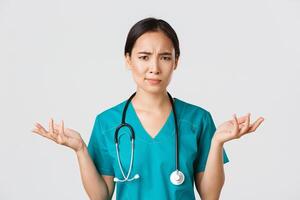 Healthcare workers, preventing virus, quarantine campaign concept. Close-up of confused and puzzled asian female physician in scrubs having complicated problem, raising hands and shrugging photo