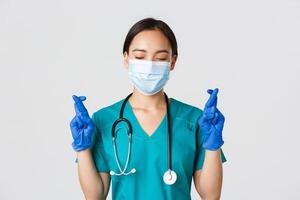 Covid-19, coronavirus disease, healthcare workers concept. Close-up of hopeful asian female doctor, physician in medical mask and rubber gloves, making wish, cross fingers good luck photo