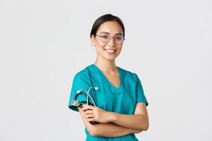 Covid-19, coronavirus disease, healthcare workers concept. Professional good-looking asian doctor, medical worker in glasses and scrubs, cross arms and smiling, white background photo