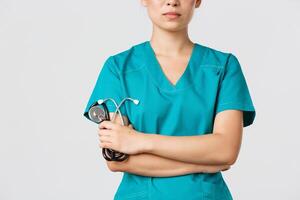 Covid-19, coronavirus disease, healthcare workers concept. Close-up of confident female asian physician, doctor in medical scrubs, cross arms determined and holding stethoscope, white background photo