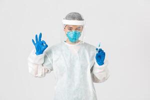 Covid-19, coronavirus disease, healthcare workers concept. Confident serious female asian doctor in personal protective equipment, show okay gesture, hold syringe with vaccine, white background photo