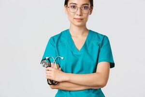 Covid-19, coronavirus disease, healthcare workers concept. Confident smiling professional asian doctor in glasses, cross arms chest, wearing scrubs and holding stethoscope, white background photo