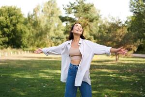 Carefree asian girl laughing and dancing in park, enjoying summer sunny day, raising hands up and breathing fresh air photo