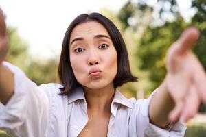 People and mobile connection. Happy young asian woman takes selfie on smartphone, holds camera with hands, poses in park on summer day photo