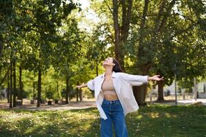 Carefree asian girl dancing, feeling happiness and joy, enjoying the sun on summer day, walking in park with green trees photo