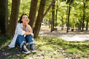 Young people. Beautiful asian girl sits near tree in park and rests, smiling and looking into distance, relaxing outdoors on fresh air in summer photo