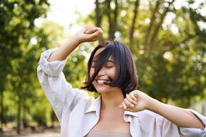 People lifestyle. Portrait of young brunette woman dancing, smiling and laughing, walking in park with hands lift up high, enjoying summer day outside photo