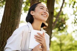 Romantic smiling girl reading book in park or foret, sitting under tree shade on sunny day, relaxing on fresh air surrounded by nature photo