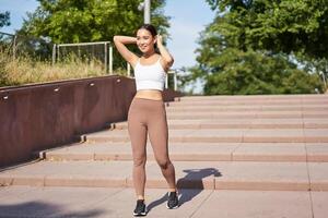 Sportswoman jogging outdoors. Smiling asian fitness girl in wireless earphones, listening music and running on road, workout photo