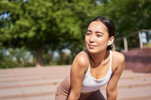 Portrait of asian woman taking break, breathing heavily and panting after running, jogger standing and wiping sweat off forehead, smiling pleased photo