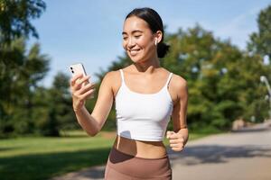 Fitness woman with water bottle and smartphone, jogging in park and smiling, looking at her mobile phone app, checking sport application photo